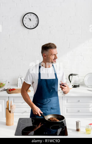 Mann in Schürze mit Glas Wein kochen Steak auf Pan Stockfoto