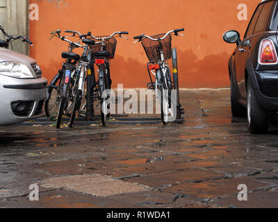 Orange Mauer gibt dramatischen Hintergrund zu vier abgestellte Fahrräder in einem fahrradträger zwischen zwei Autos auf einer nassen Fahrbahn in Florenz, Toskana, Italien Stockfoto