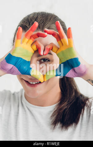 Junge transgender Mann an der Kamera schaut, während sie Herz mit Hände in Farben von Stolz Flagge vor der weißen Wand Stockfoto