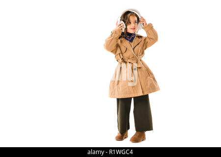 Schöne kleine Kind in Trenchcoat mit Kopfhörern isoliert auf weißem Stockfoto