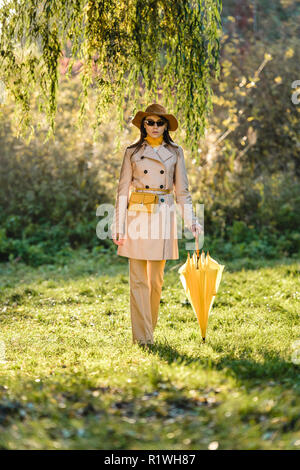 Elegante junge Frau mit Sonnenbrille, Trenchcoat und Hut mit yellow umbrella posiert auf der Wiese Stockfoto