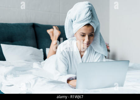 Schöne junge Frau im Bademantel mit Laptop beim liegen auf dem Bett am Morgen Stockfoto