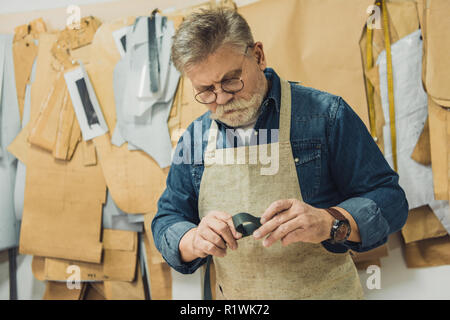 Konzentriert Männer mittleren Alters Handtasche Handwerker in Schürze und Brillen arbeiten mit Stoff im Studio Stockfoto