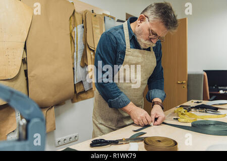 Selektiver Fokus der Männer mittleren Alters Handtasche Handwerker in Schürze und Brillen arbeiten mit Stoff im Studio Stockfoto