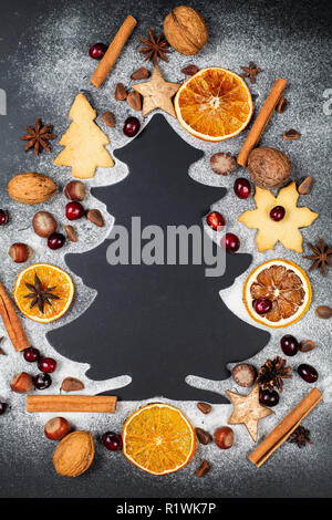 Form der Weihnachtsbaum aus Puderzucker auf dunklem Hintergrund mit Muttern, Preiselbeeren, Sternanis, Pinienkerne, Cookies und trockenen Scheiben von Orange Stockfoto