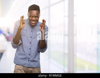 Junge afrikanische amerikanische Mann mit blauem Hemd lächelnd die Daumen mit Hoffnung und die Augen geschlossen. Glück und abergläubischen Konzept. Stockfoto