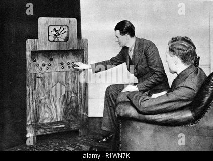 Ein 1935 Foto, die frühen Tage des Fernsehens in Großbritannien - zwei Männer in Anzügen sitzen zu sehen Micky Maus spielt Dudelsack auf dem kleinen Fernseher in einem großen Holzgehäuse untergebracht Stockfoto