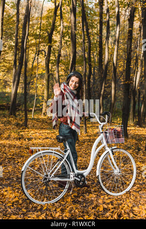 Lächelnd stilvolle Mädchen in Lederjacke und Baskenmütze winkte mit der Hand und mit dem Fahrrad im herbstlichen Park Stockfoto