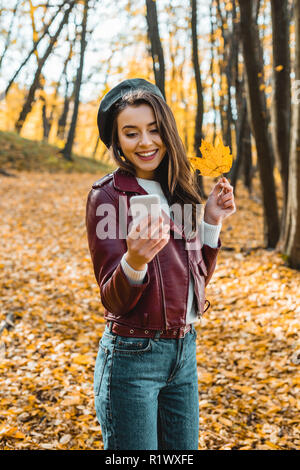 Lächelnd stilvolle Mädchen in Lederjacke unter selfie mit gelben Blatt auf dem Smartphone im Freien Stockfoto