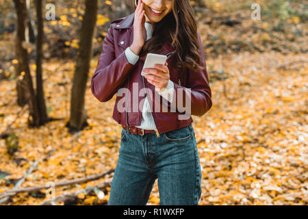 Zugeschnittenes Bild der lächelnde Frau in stilvollem Leder Jacke mit Smartphone im herbstlichen Wald Stockfoto
