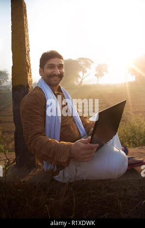 Bauer sitzt in der Nähe von seiner Landwirtschaft Feld mit einem Laptop und lächelnd. Stockfoto