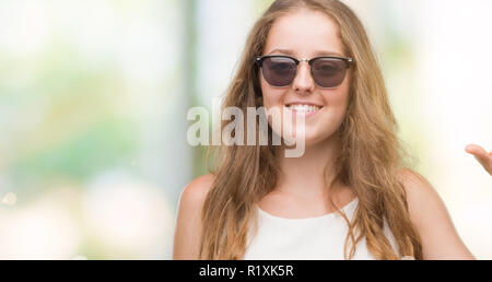 Junge blonde Frau mit Sonnenbrille lächelnd und selbstbewusst und deutete mit der Hand tun Größe Schild mit Finger, während sie und die Kamera. Messen con Stockfoto