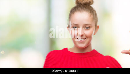 Junge blonde Frau mit Brötchen und roten Pullover lächelnd und selbstbewusst und deutete mit der Hand tun Größe Schild mit Finger, während sie und die Kamera. Mir Stockfoto