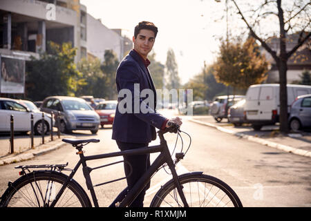Ein junger hübscher Mann lächelnd crossing Street, drücken Fahrrad im Freien, belebten Straße Autos (Unscharf im Hintergrund). Stockfoto