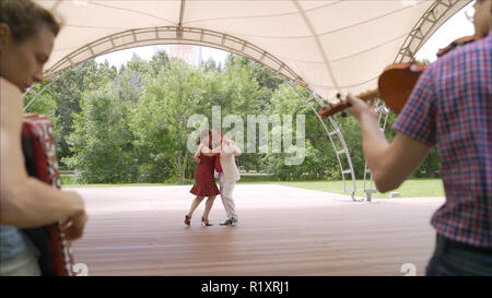 Akkordeon und Geige spielen im Park. Eine Frau in einem roten Kleid und ein Mann in einem weißen Anzug sind Tango tanzen. Ein älteres Paar Tango tanzen im Park. Stockfoto