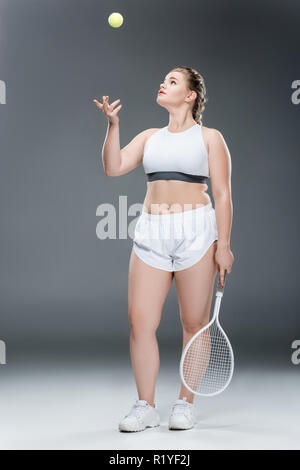 Junge übergewichtige Frau mit Tennisschläger und Ball werfen auf Grau Stockfoto