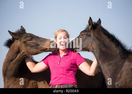 Warmblut. Lachende Frau mit verspielten Fohlen auf der Weide. Deutschland Stockfoto