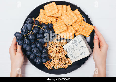 Woman's Hände halten Platte mit Käse, Trauben, Kekse und Nüsse, Ansicht von oben auf die weiße Holztisch. Stockfoto