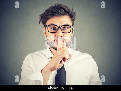 Junge hipster Mann in Brillen halten den Finger auf die Lippen, für Stille und lächelnd an der Kamera auf grauem Hintergrund Stockfoto