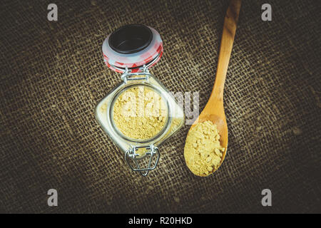 Blick von oben auf die pulverförmigen Senfkörner, gelb Senfpulver in süßen vintage Glas Glas auf rustikalen Sack dunkel braunen Hintergrund. Stockfoto