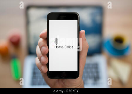 Ein Mann schaut auf seinem iPhone. das Home Office Logo, während saß an seinem Computer Schreibtisch (nur redaktionelle Nutzung). Stockfoto