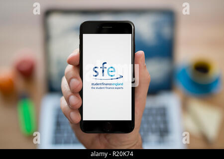 Ein Mann schaut auf seinem iPhone zeigt der Student Finance England (SFE) Logo, während saß an seinem Computer Schreibtisch (nur redaktionelle Nutzung). Stockfoto