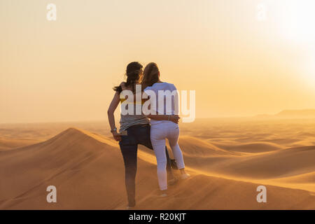 Paar junge touristische Frauen bei Sonnenuntergang in Sanddünen in Dubai Wüste suchen. Schöne Aussicht auf die Wüste und den Sonnenuntergang von der Oberseite der Sanddünen Stockfoto