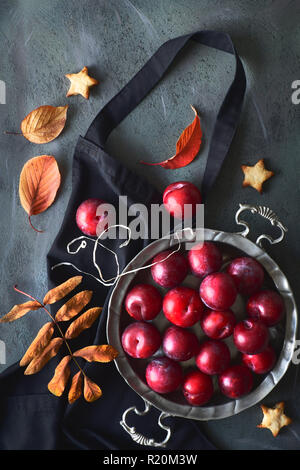 Vintage Metallplatte mit rote Pflaumen, Star cookies und verstreut rot und orange Herbst Blätter auf schwarze Schürze auf dunklem Hintergrund Stockfoto