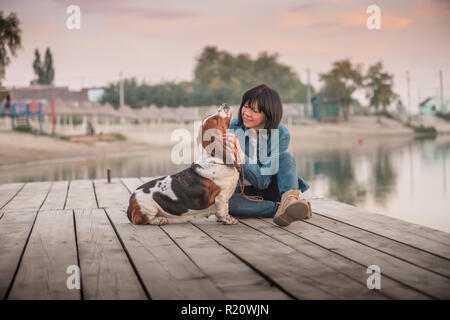 Porträt der schönen jungen Frau spielen mit Hund am Fluss. Glückliche Frau sitzen auf dem Holzsteg mit ihrem Hund Basset Hound. Frau mit Welpen. Stockfoto