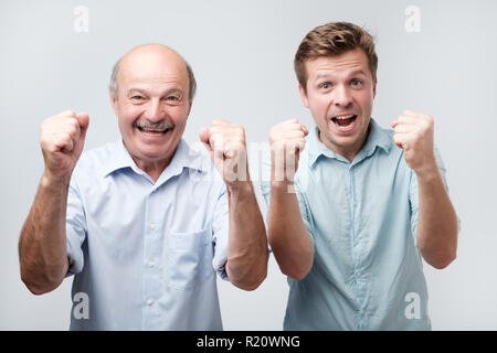 Foto der freudigen zwei junge Männer clench Fists und Schreien mit Glück, gekleidet wie nebenbei, auf weißem Hintergrund. Happy Sohn und Vater feiern Stockfoto