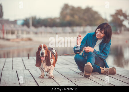 Porträt der schönen jungen Frau spielen mit Hund am Fluss. Glückliche Frau sitzen auf dem Holzsteg mit ihrem Hund Basset Hound. Frau mit Welpen. Stockfoto