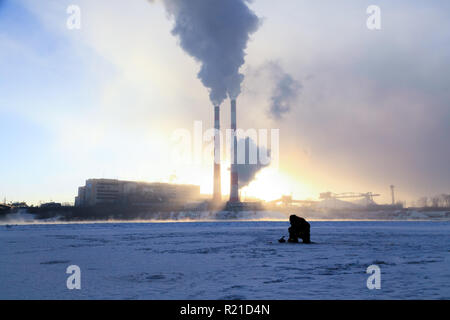 Winter angeln, Leidenschaft, Fischer fangen Fische auf einem zugefrorenen Fluss vor dem Hintergrund der Fabrik Rohre bei Sonnenuntergang. Konzept von Öl und Gas pr Stockfoto