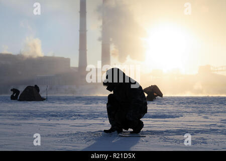 Winter angeln, Leidenschaft, Fischer fangen Fische auf einem zugefrorenen Fluss vor dem Hintergrund der Fabrik Rohre bei Sonnenuntergang. Konzept von Öl und Gas pr Stockfoto