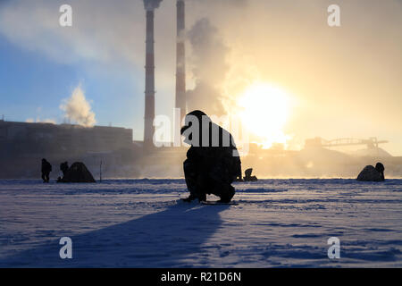 Winter angeln, Leidenschaft, Fischer fangen Fische auf einem zugefrorenen Fluss vor dem Hintergrund der Fabrik Rohre bei Sonnenuntergang. Konzept von Öl und Gas pr Stockfoto