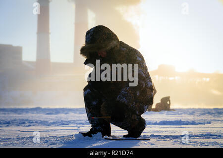 Winter angeln, Leidenschaft, Fischer fangen Fische auf einem zugefrorenen Fluss vor dem Hintergrund der Fabrik Rohre bei Sonnenuntergang. Konzept von Öl und Gas pr Stockfoto