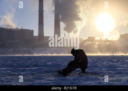 Winter angeln, Leidenschaft, Fischer fangen Fische auf einem zugefrorenen Fluss vor dem Hintergrund der Fabrik Rohre bei Sonnenuntergang. Konzept von Öl und Gas pr Stockfoto