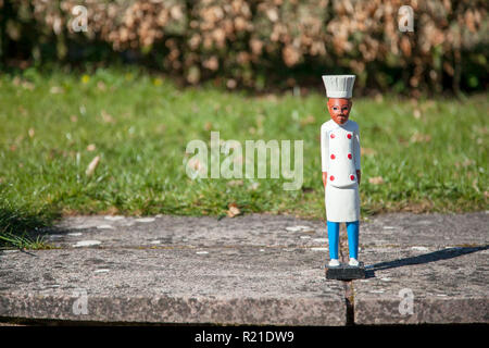 Kleine Holzfigur eines dunkelhäutigen Koch trägt einen weißen Mantel und Hut, stehend auf Pflastersteinen vor einer Rasenfläche, auf der Rasenfläche. Stockfoto