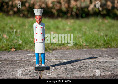 Kleine Holzfigur eines dunkelhäutigen Koch trägt einen weißen Mantel und Hut, stehend auf Pflastersteinen vor einer Rasenfläche, auf der Rasenfläche. Stockfoto