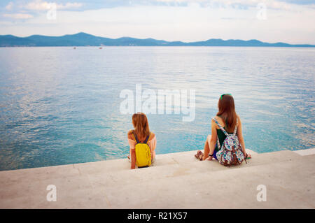 Zwei junge Mädchen mit Rucksäcken auf der Steintreppe in der Nähe der Küste mit Blick auf die Hügel in der Ferne. Stadt Zadar, Kroatien Stockfoto
