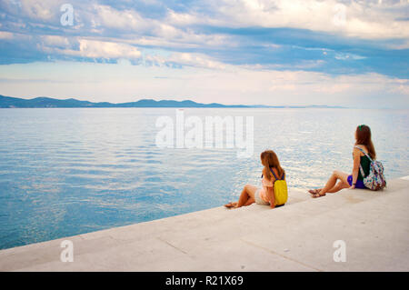 Zwei junge Mädchen mit Rucksäcken auf der Steintreppe in der Nähe der Küste suchen wehmütig auf die Hügel in der Ferne und entspannend. Zadar, Stockfoto
