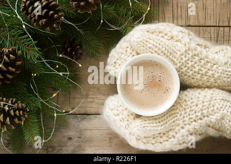 Frauen Hände in Handschuhe Hug eine Tasse Kakao auf dem Hintergrund von tannenzweigen mit Tannenzapfen und eine Girlande. Selektive konzentrieren. Stockfoto