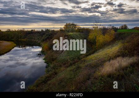 Ruhigen schmalen Flusses von seiner steilen Bank gesehen, mit grünem Gras und Büschen bedeckt. Bunte Herbst Abend Landschaft mit der Landschaft, mit Bewölkt Stockfoto