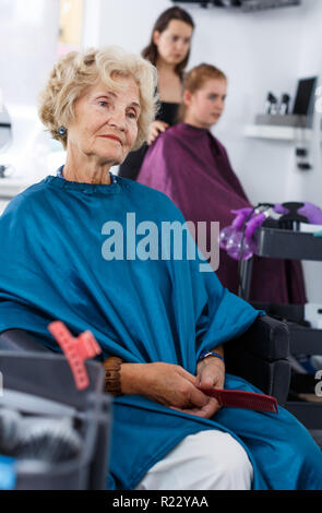 Elegante ältere Frau in Friseursalons Cape in Friseur Stuhl Sitzen, Warten auf Restaurantbesuche Stockfoto