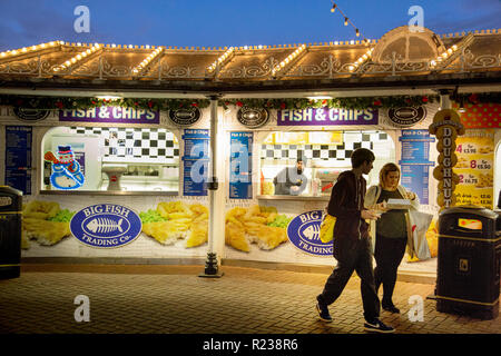 Auf Fisch-und-Chip auf Brighton Pier ein junger Mann und eine Frau Essen kaufen. Overhead, das Dach hat Zeilen von Glühbirnen unter einem Abendhimmel. Stockfoto