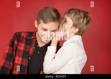 Junge Frau flüstert das Geheimnis zu lächeln Ohr des Menschen. Studio auf rotem Hintergrund Stockfoto