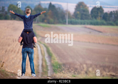 Rückansicht des athletischen Vater Tragen auf den Schultern Sohn Spaziergang durch Wiese auf verschwommenen nebligen grünen Bäumen und blauer Himmel. Aktives Leben Stockfoto