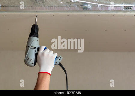 Nahaufnahme der Konstruktor Arbeitnehmer Hand in Schutzhandschuh mit elektrischer Akkuschrauber Anschluss ausgesetzt Trockenbau decke Metallrahmen. Reno Stockfoto