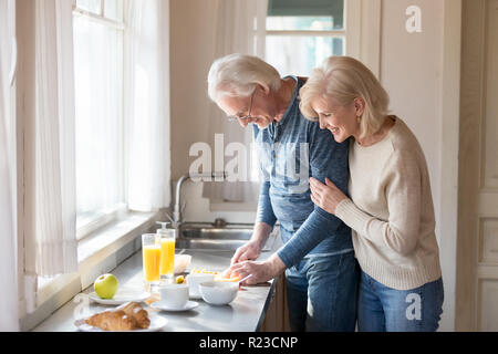 Lächelnd im Alter von Frau hug Mann hinter ihn aufpassend, Zubereitung von gesunder Nahrung, liebevolle ältere Frau umarmen Mann kochen Frühstück schneiden Obst, Romant Stockfoto