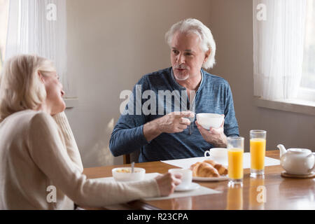 Gerne älteres Paar genießen morgens zu Hause essen gesund, Frühstück, Kaffee, lächelnd im Alter von Mann und Frau sprechen am Küchentisch Ausgaben weeken Stockfoto