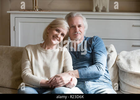 Portrait von lächelnden im Alter von Mann und Frau sitzen auf der Couch zu Hause halten sich an den Händen posieren für Bild, Senior Paar Umarmung entspannt auf einem Sofa am Cam suchen Stockfoto